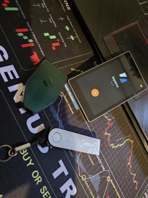 A photograph of cryptocurrency hardware wallets and a Kraken branded card reader resting on a desk mat printed with financial charts and candlestick patterns.