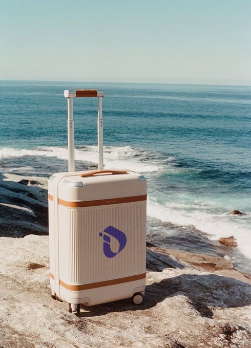 A cream-colored rolling suitcase with an orange stripe sits on coastal rocks overlooking the ocean, branded with a blue 'D' logo.