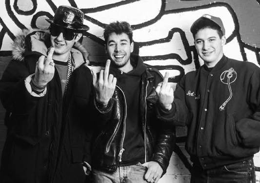 A black and white photo shows three young men in jackets smiling and making middle finger gestures while standing against a graffiti-covered wall.