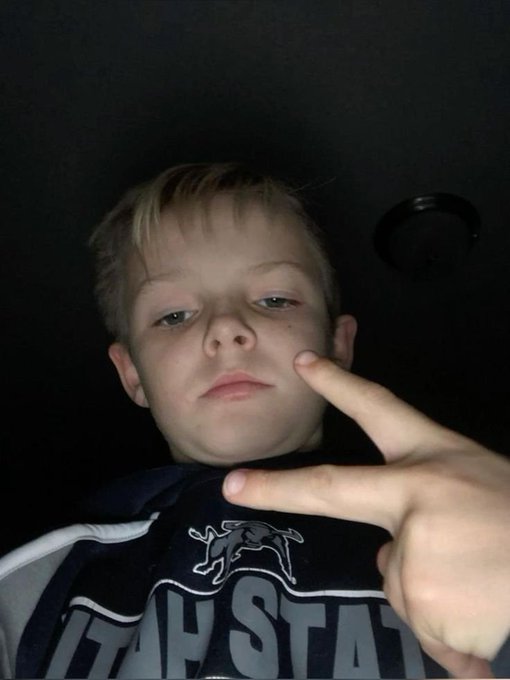 A young boy with blond hair posing for a selfie, making a hand gesture near his face while wearing a dark blue shirt with a horse logo and the word 'STATE'.