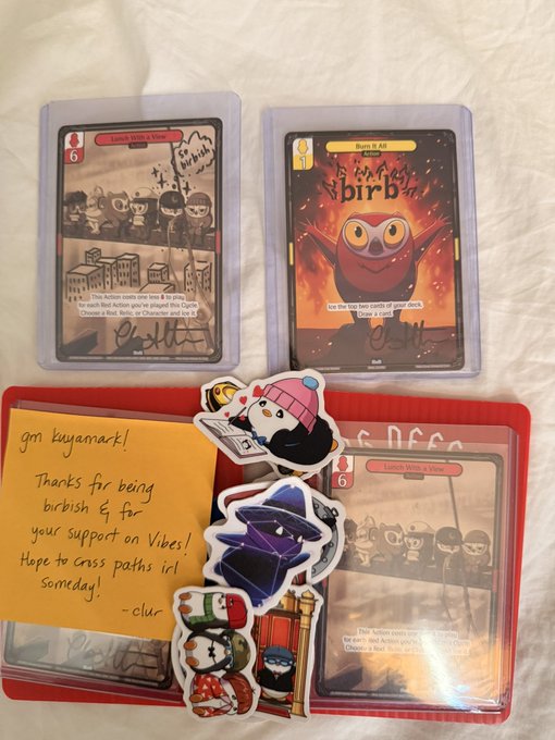 A collection of trading cards, stickers, and a handwritten note on orange paper that says 'gm bayamck! Thanks for being birbish & for your support on Vibes! Hope to cross paths irl someday! - clur -'.