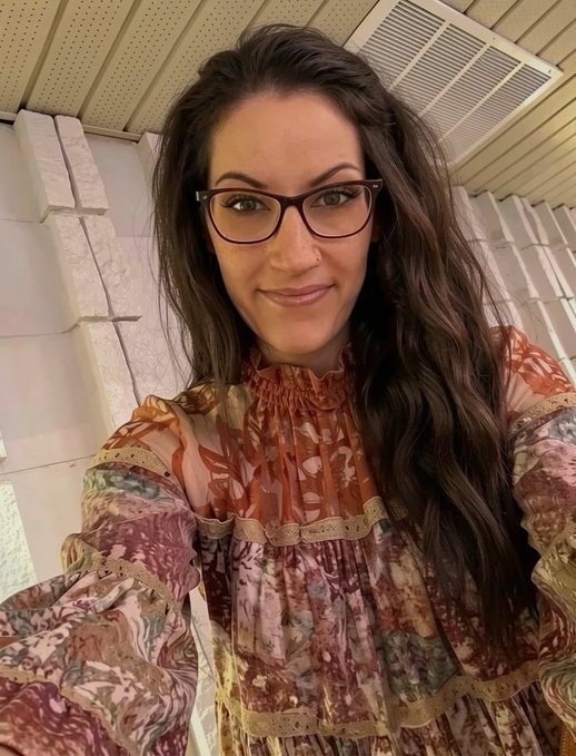 A selfie of a smiling woman with long dark hair and black-rimmed glasses, wearing a ruffled, earth-toned patterned blouse.