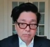 A man with dark hair and thick-rimmed glasses wearing a dark suit jacket and white shirt, photographed against a light background.
