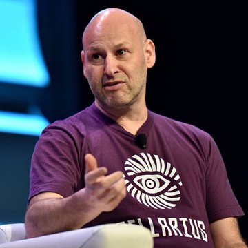 A bald man in a purple "ILLARIUS" t-shirt with an eye logo gestures with his hand while speaking. The image is referenced as Joseph Lubin, CEO of ConsenSys and early Ethereum funder, though his appearance here is typical of his public speaking engagements.