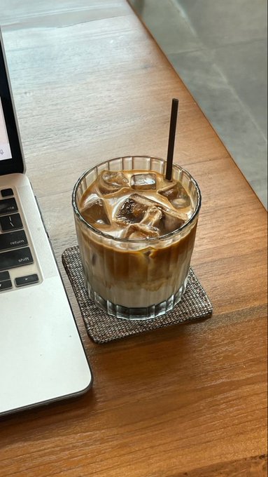 An iced coffee drink in a glass with a straw, sitting on a coaster next to a laptop on a wooden table.