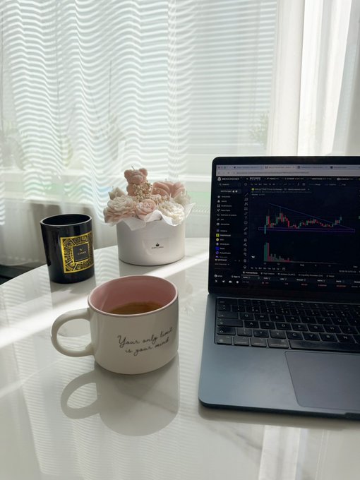 A cozy desk setup features a laptop displaying a stock chart, a candle in a white container, a black and yellow candle, and a white mug reading 'Your only limit is your mind' filled with a beverage.