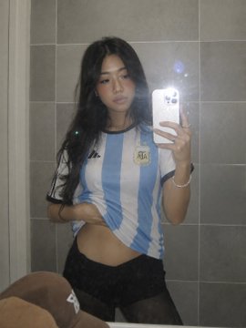 A mirror selfie of a woman with long dark hair wearing an Argentina national football jersey (light blue and white stripes with AFA crest) and black shorts, holding a smartphone in a tiled bathroom.