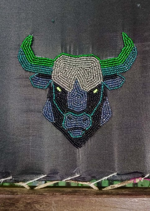 A beaded embroidery artwork depicting a stylized bull or minotaur head with green horns and blue accents on a dark gray fabric background.