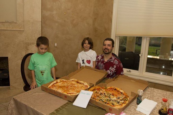 A family photo featuring two children and a man sitting at a table with two large pizzas in open boxes, one of which is labeled 'PMI A02', with a bottle of water visible on the table.