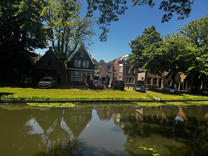 A scenic view of a canal reflecting traditional Dutch-style houses and lush green trees under a clear blue sky.