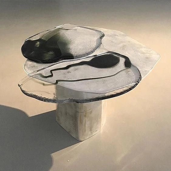 An abstract sculptural piece featuring a translucent, irregularly shaped top with black organic forms and flowing lines, mounted on a solid white cubic pedestal. The artwork casts a soft shadow on the neutral surface beneath it.