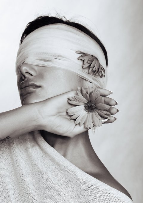 A black-and-white photograph of a person with a bandage covering their eyes, holding a sunflower over their mouth.