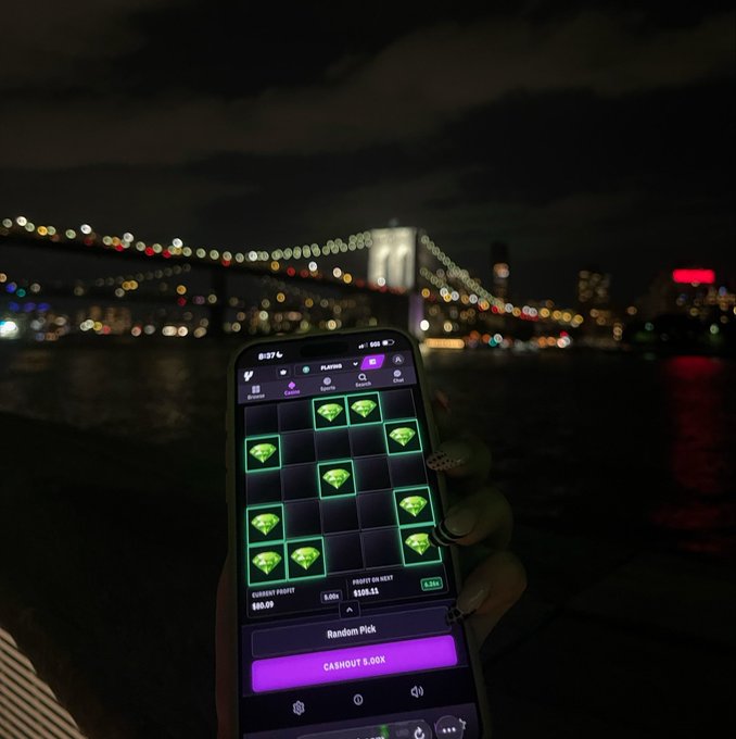 A person holds up a smartphone displaying a crypto casino-style game with green diamond tiles, a "$50.99" current points balance, a "$128.13" profit on next, and a "CASHOUT 0.00X" button, set against the nighttime backdrop of the illuminated Brooklyn Bridge and city skyline. The image visually reinforces the post's message of stepping away from the exhausting grind of memecoin trading, replacing high-stakes crypto speculation with a casual mobile gaming break by the waterfront.