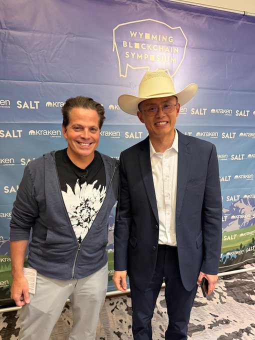 Two men smile together at the Wyoming Blockchain Symposium, posing in front of a step-and-repeat banner featuring "SALT" and "Kraken" sponsor logos, with one wearing a cowboy hat to match the Wyoming theme of the post.