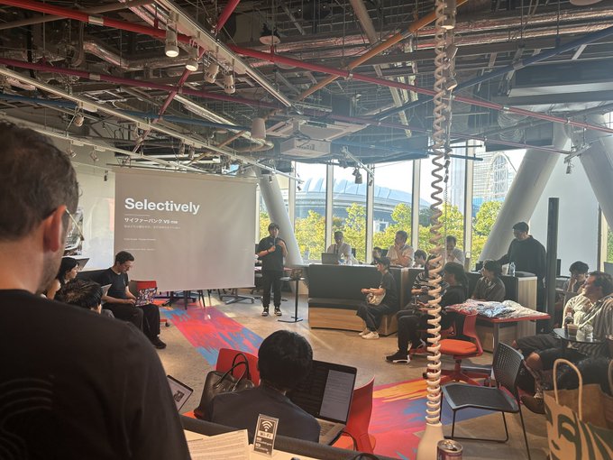 A photograph of a presentation in a modern, industrial-style office space, with a speaker standing before a projection displaying the word 'Selectively' while an audience sits on couches and chairs.