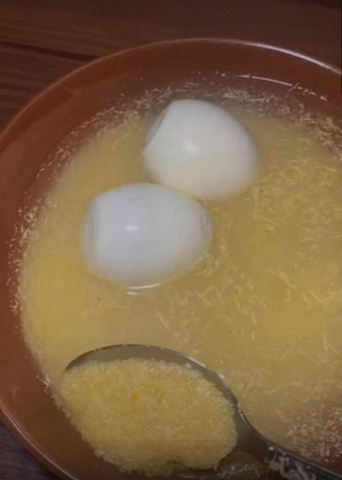 A bowl of yellow soup or broth containing two white round dumplings, with a spoon resting in the bowl.