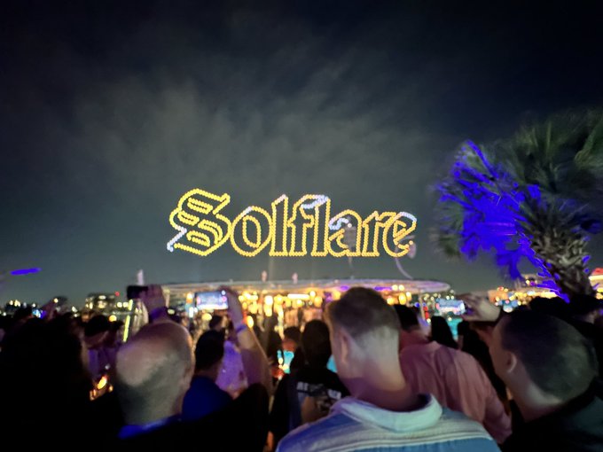 A nighttime photo of a crowd at an event with a large illuminated sign reading 'Solflare' in cursive yellow lights, with palm trees and festival lights visible in the background.