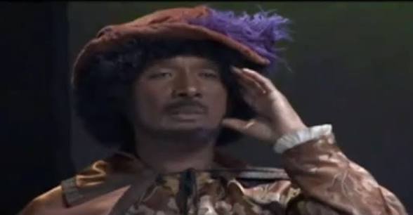 A still of a man in a feathered hat and ornate period clothing holding his hand to his ear, as if listening.