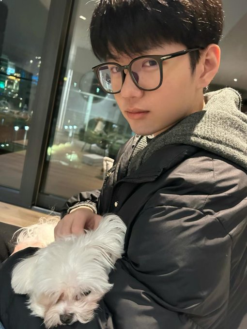 A young man wearing glasses and a dark winter jacket holds a small white dog in his arms while taking a selfie indoors at night.