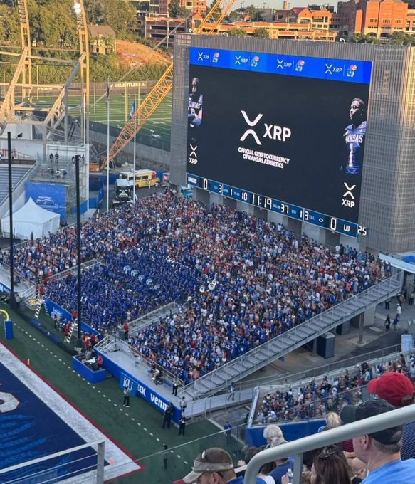 Stadium crowd photo showing the jumbotron displaying XRP crypto logo with text 'OFFICIAL CRYPTOCURRENCY OF KANSAS JAYHAWKS' above a scoreboard showing KU 19, LW 0.