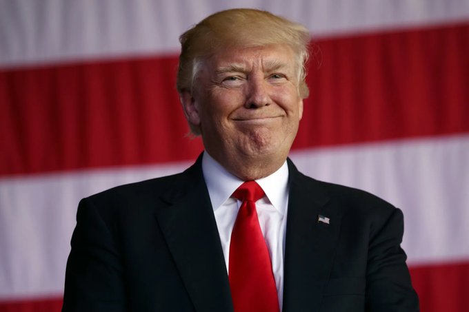 Donald Trump smiles slightly while wearing a dark suit and red tie, standing in front of an American flag background.