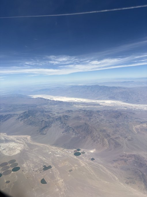 An aerial view from an airplane window shows a vast desert landscape with mountains, a salt flat or dry lake bed, and circular agricultural irrigation patterns in the valley below.