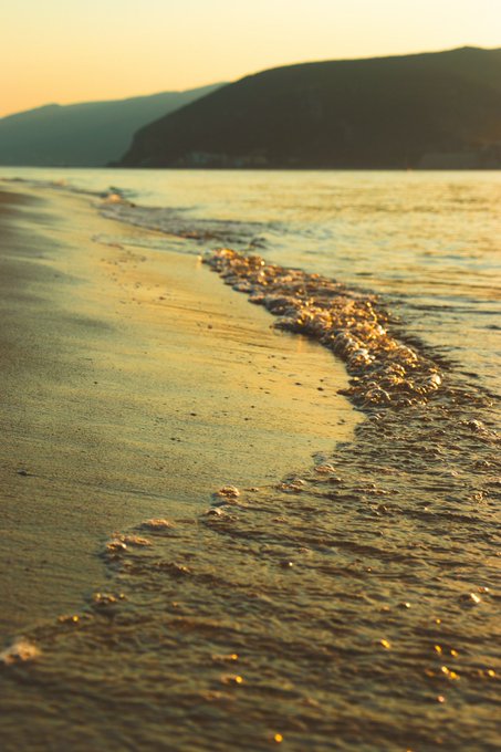 A sunset view of gentle waves washing onto a sandy beach, with a dark mountainous coastline visible in the background.