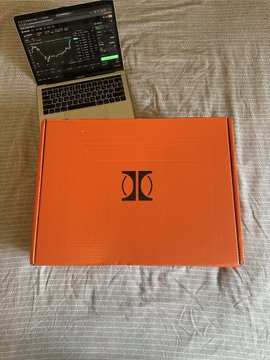 An orange shoe box with a black logo sits on a bed next to a laptop displaying trading charts with green indicators.