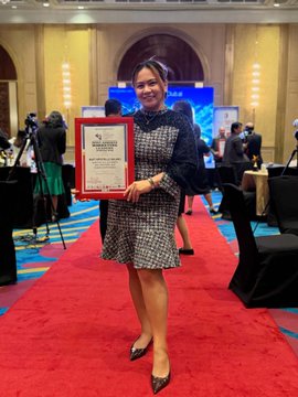 A smiling woman stands on a red carpet in a patterned dress, proudly holding a framed certificate from SheFi as a Scholar selection for Season 17.