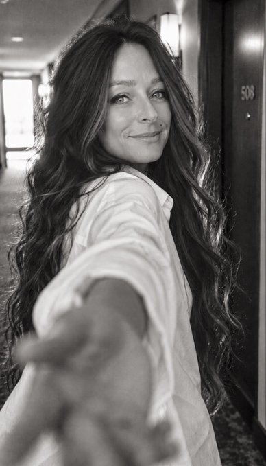 A black-and-white portrait of a woman with long wavy hair, wearing a white shirt, smiling and reaching out her hand toward the camera in a hallway.