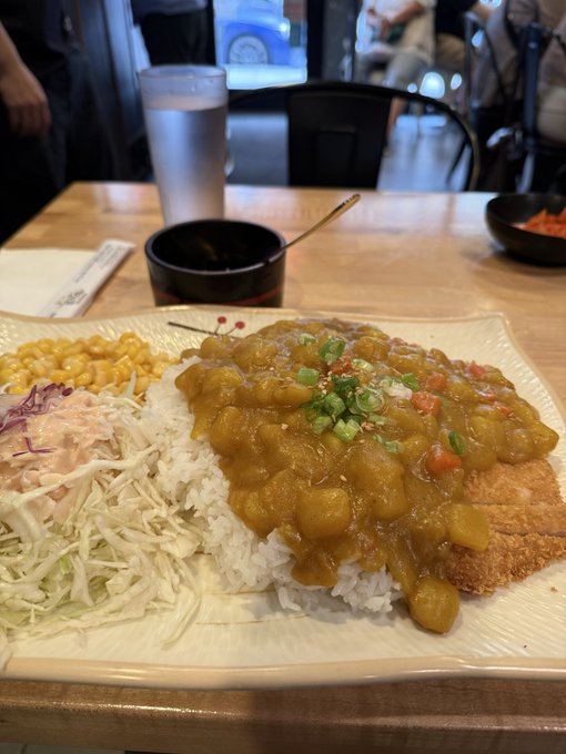 A Japanese curry rice dish with a breaded cutlet, topped with diced onions and scallions, served alongside corn and shredded cabbage at a restaurant table.