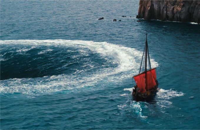 A scenic photograph of a small sailboat with red sails navigating turbulent blue ocean waters near a rocky cliff.
