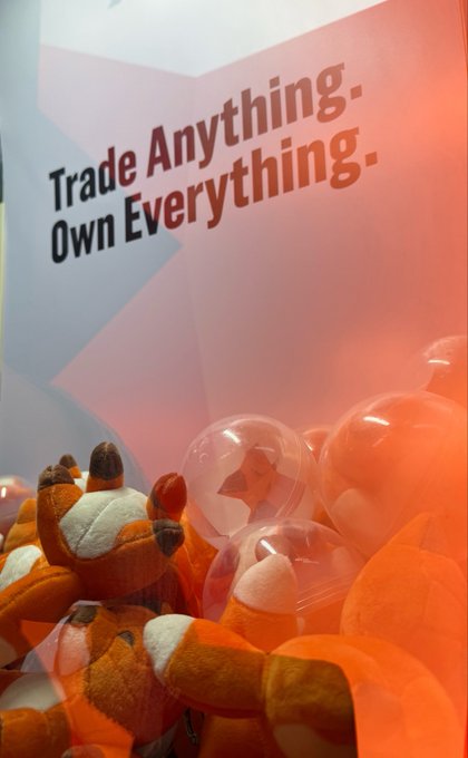 A claw machine filled with orange fox plush toys features a sign reading 'Trade Anything. Own Everything.' in the background.