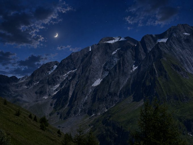 A scenic night landscape featuring rugged, rocky mountains under a dark blue sky with scattered clouds, a crescent moon, and visible stars, with a forested valley in the foreground.