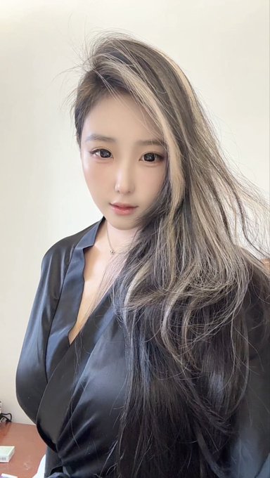 A selfie of a young woman with long highlighted hair wearing a black satin blouse and a delicate necklace.