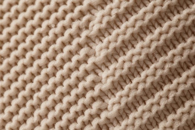 A close-up of a cream-colored knitted textile with a visible diagonal weave pattern of interlocking yarn loops, exemplifying the tactile, yarn-based textures characteristic of NEO's collection.