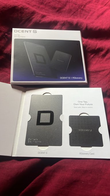 A newly unboxed D'CENT S hardware wallet and R3covery card sit in their packaging on a red fabric surface, displaying the "DCENT S x R3covery" branding and "One Tap, Own Your Future" tagline, complementing the post announcing the user's new crypto storage purchase.