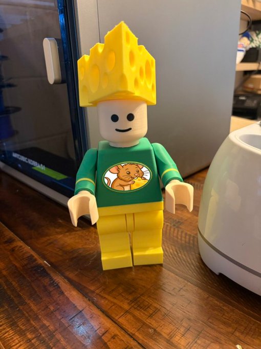 A small LEGO-style figure wearing a yellow cheese-shaped crown and a green shirt with a lion graphic sits on a wooden table next to a white appliance.