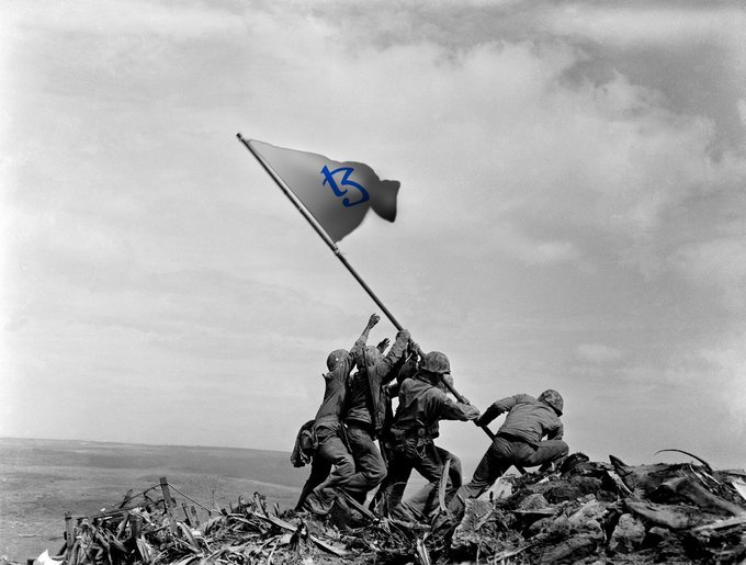 Soldiers raise a flag with a blue symbol on a debris-strewn hill, mimicking the iconic Iwo Jima flag-raising photo, with the text "Dying on the tezos hill!" indicating a cryptocurrency context.