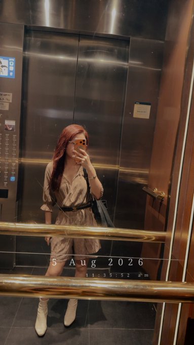 A mirror selfie of a woman with red hair in a beige dress and white boots taken inside an elevator, dated 5 Aug 2026.