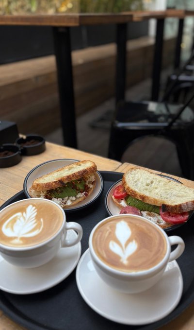Two cappuccinos with leaf latte art and two sandwiches on a black tray at a cafe table, with a wooden table and black chair in the background.