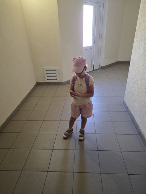 A young child wearing a pink cap, yellow t-shirt with a small design, pink shorts, sandals, and a backpack stands on a tiled hallway floor, looking down at a smartphone held in both hands.
