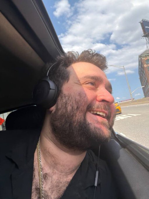 A bearded man wearing JBL headphones, a black blazer, and a gold chain smiles while sitting in a car with a city skyline visible outside.