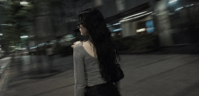 The image shows a woman walking down a street at night with her hair pulled back into a ponytail. She appears to be looking off into the distance or lost in thought as she walks. The photo has been edited, giving it a grainy appearance that adds an artistic touch and makes the scene feel more immersive.