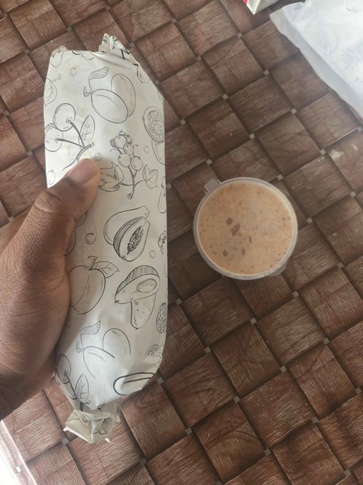 A hand holding a food wrap in decorative paper featuring fruit and botanical line drawings, alongside a plastic cup of frothy brown beverage, resting on a woven brown mat.