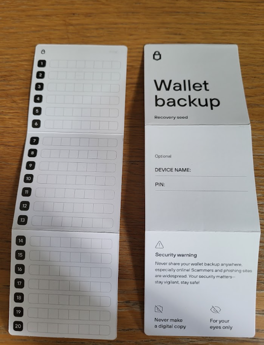 A cryptocurrency hardware wallet backup sheet is displayed on a wooden surface, featuring 20 numbered blank fields for recording a recovery seed phrase, along with instructions for an optional device name and PIN, plus security warnings.