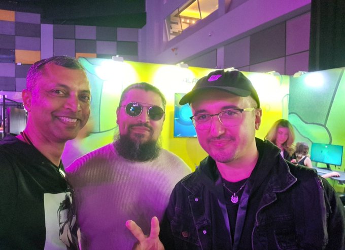 Three men posing together indoors at what appears to be an event, including one with a beard wearing sunglasses and another wearing a black cap and denim jacket, with bright yellow lighting in the background.