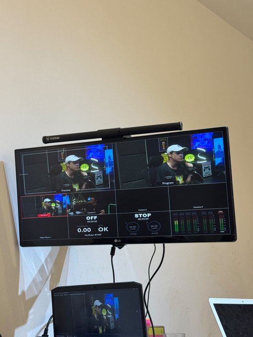 A wall-mounted LG monitor displaying a multi-camera production setup showing a man with a headset microphone, with a laptop visible below on a desk.