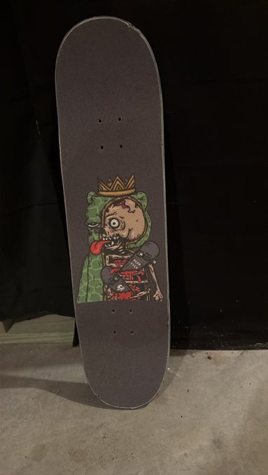 A black skateboard deck stands upright, featuring a cartoon graphic of a crowned zombie-like character with a bloody mouth and exposed ribs leaning against a green couch.