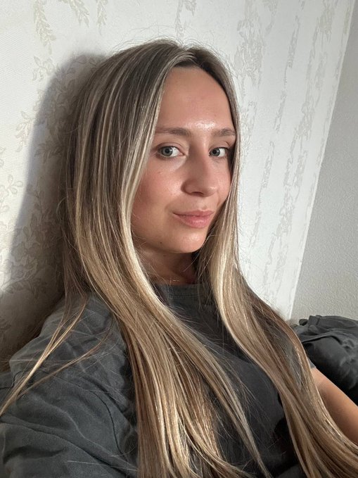A selfie-style portrait of a young woman with long blonde-highlighted brown hair, wearing a gray shirt and leaning against a floral-patterned wall.
